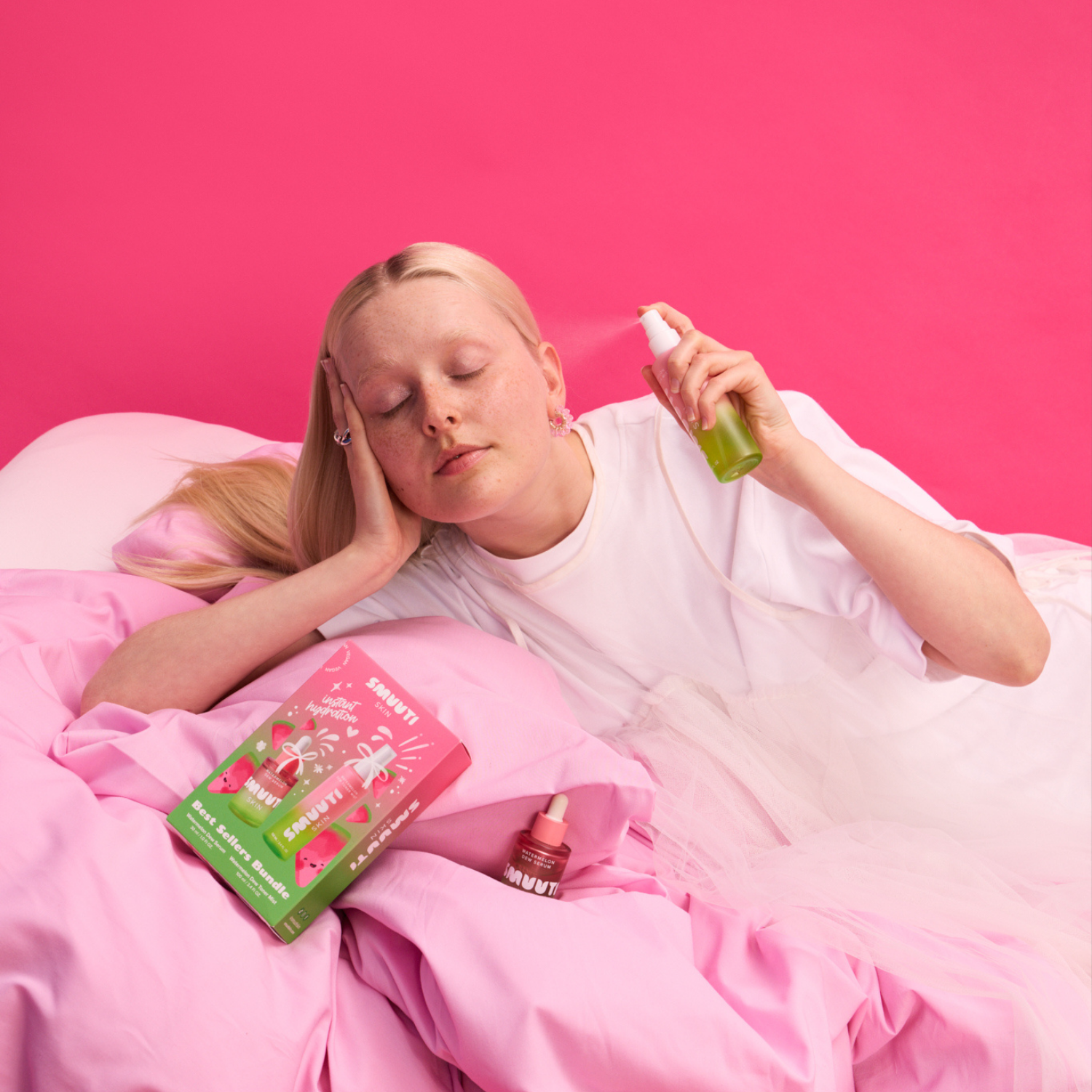 A woman lying in bed with pink bedding and a bright pink background, holding a watermelon dew toner mist. Best seller bundle is lying in the bed