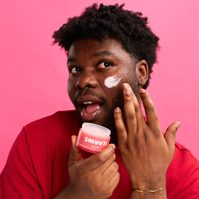 A man with dark curly hair applies white cream to his cheek while holding a pink jar of Smuuti Skin strawberry boost capsule cream. He is wearing a red shirt and posing against a solid pink background.