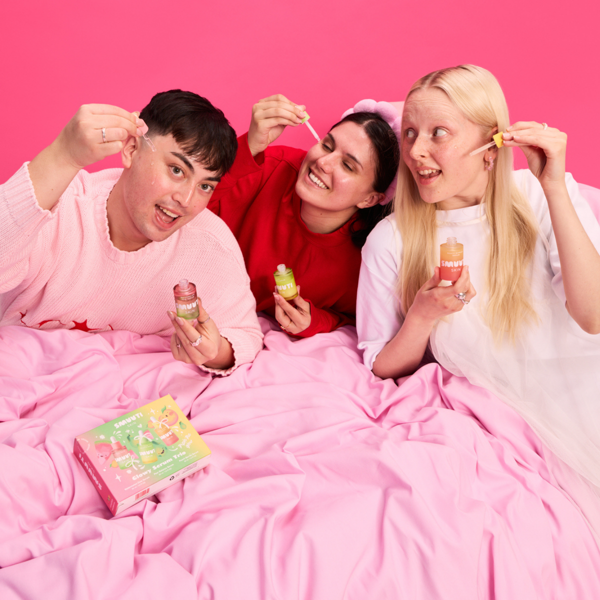 Three people lying in a bed covered with pink sheets, holding Smuuti Skin serums