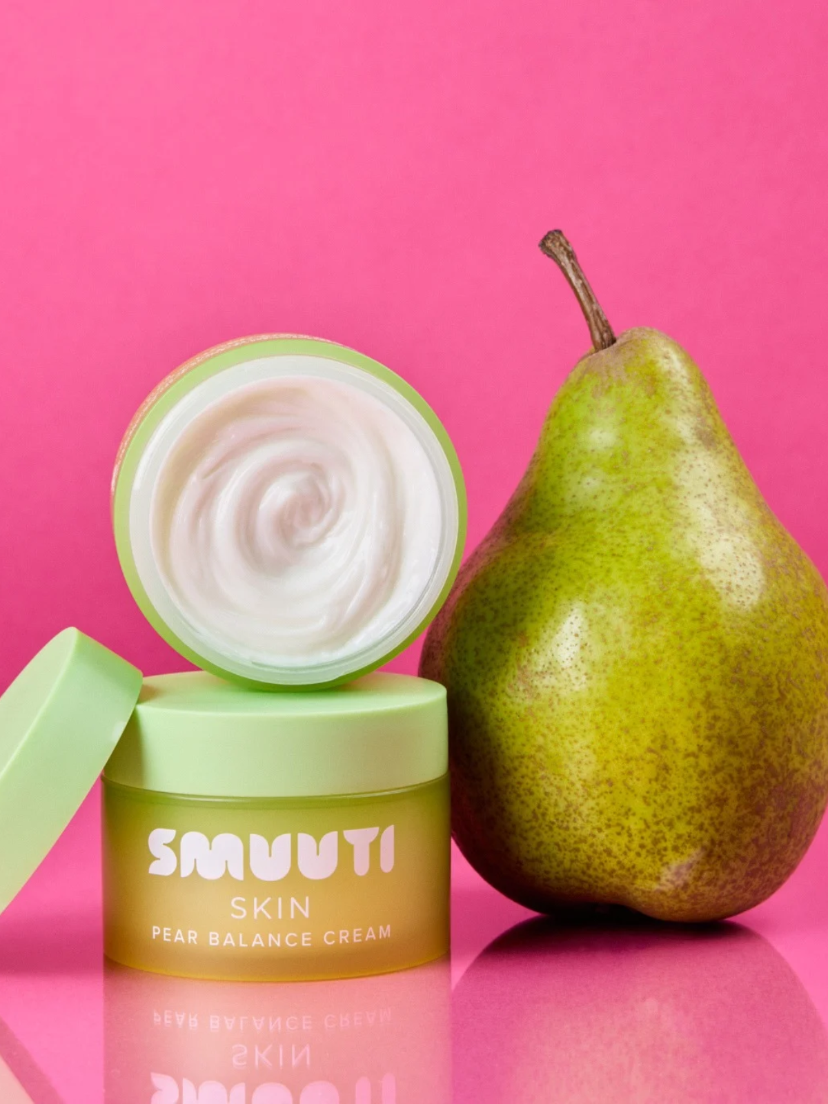 An open jar of SMUUTI Skin Pear Balance Cream with white cream visible inside, placed next to a whole pear on a bright pink reflective surface.