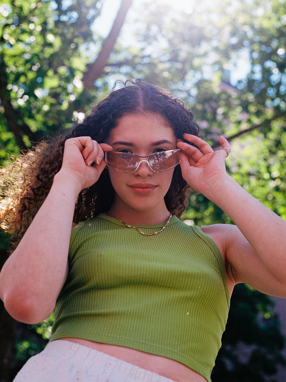 Woman wearing sunglasses with a green top and white pants against a blurred natural background
