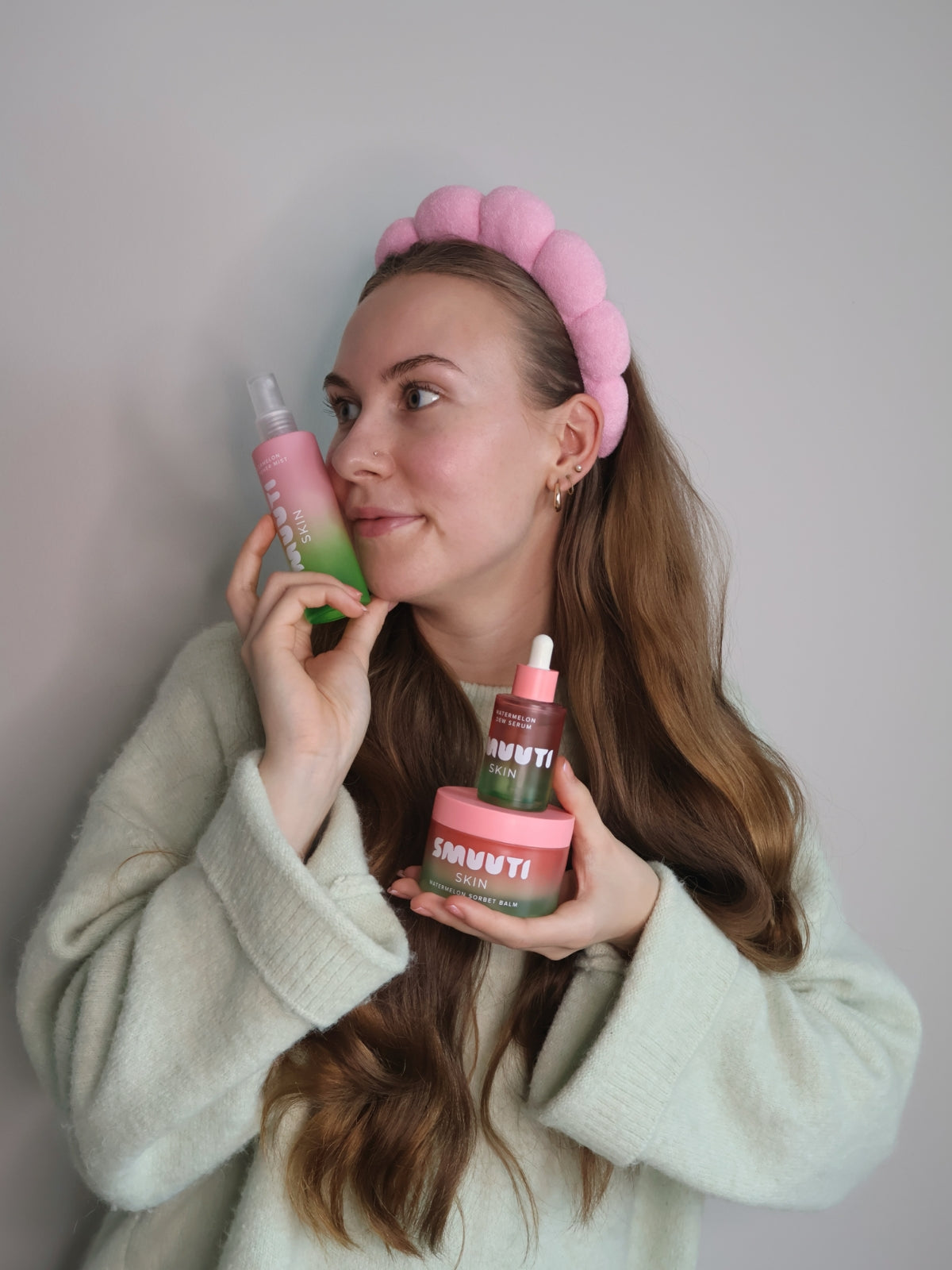 Woman with a pink skincare headband holding and posing with smuuti skin skincare products against a plain background