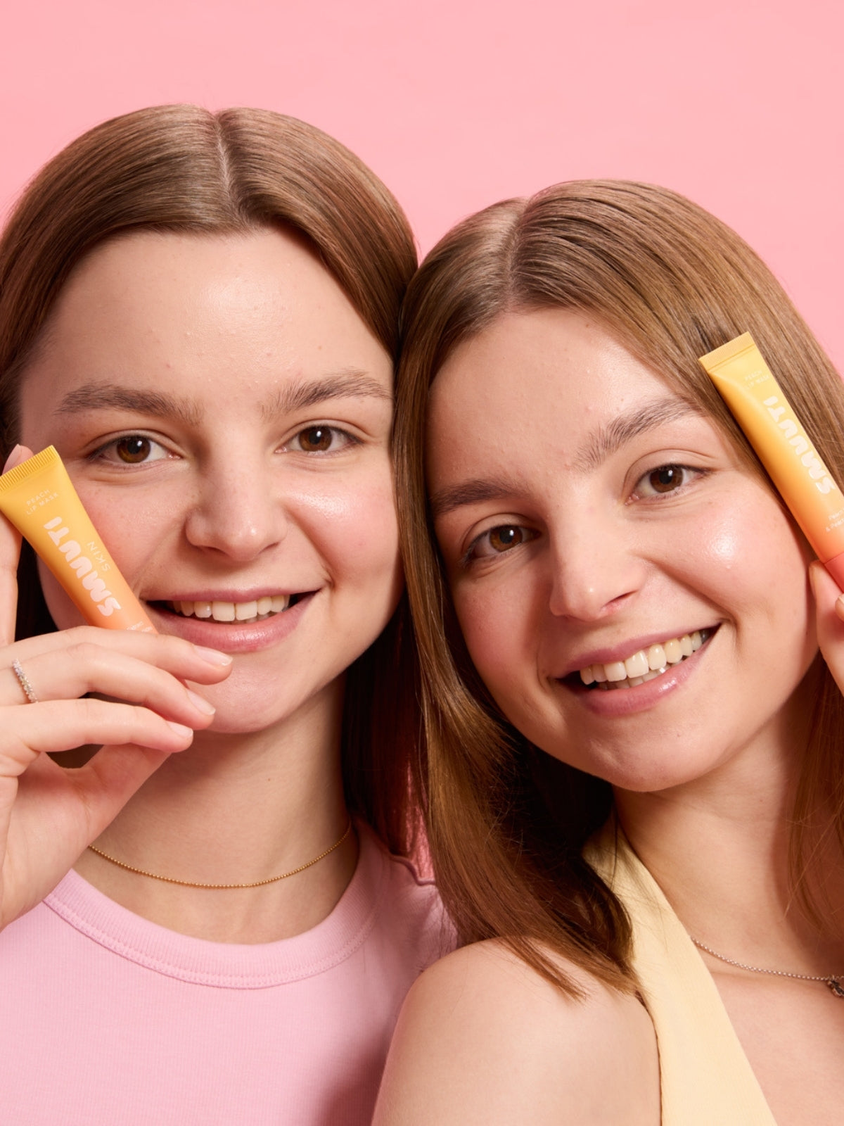 Two women holding Smuuti Skin lip masks against a pink background