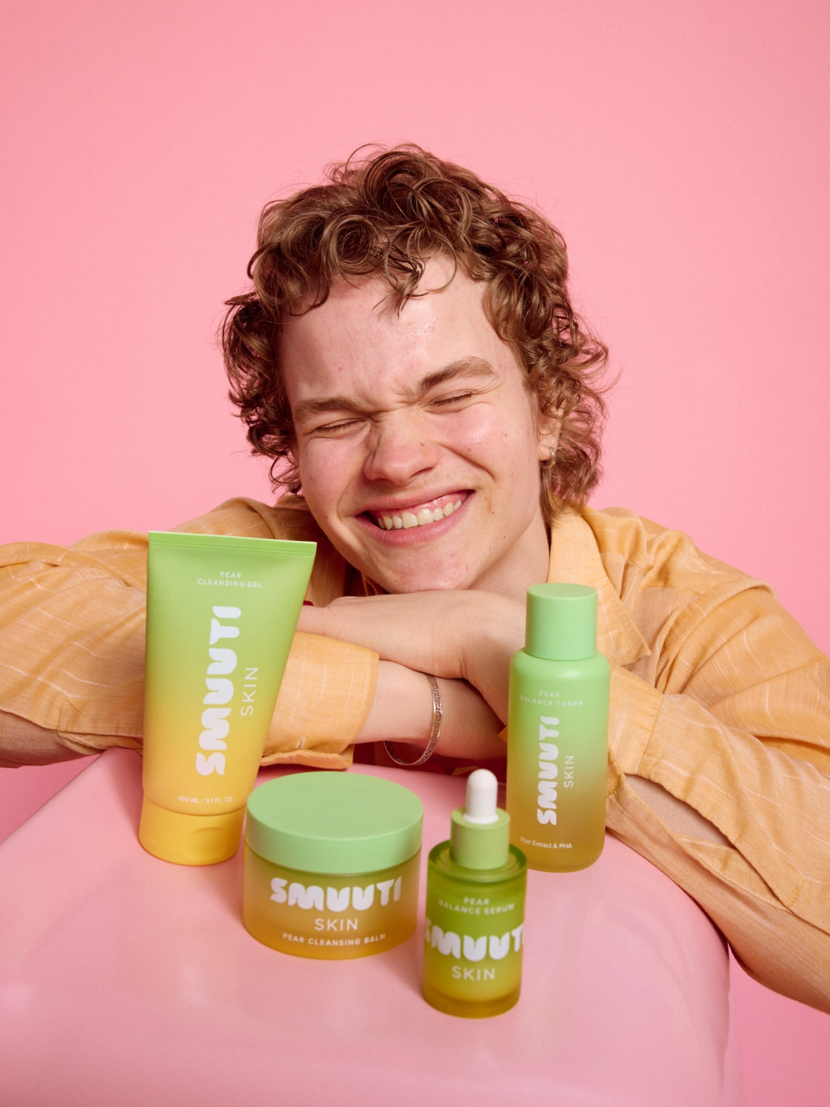 Person with curly hair sitting behind Smuuti Skin pear balance skincare products on a pink background