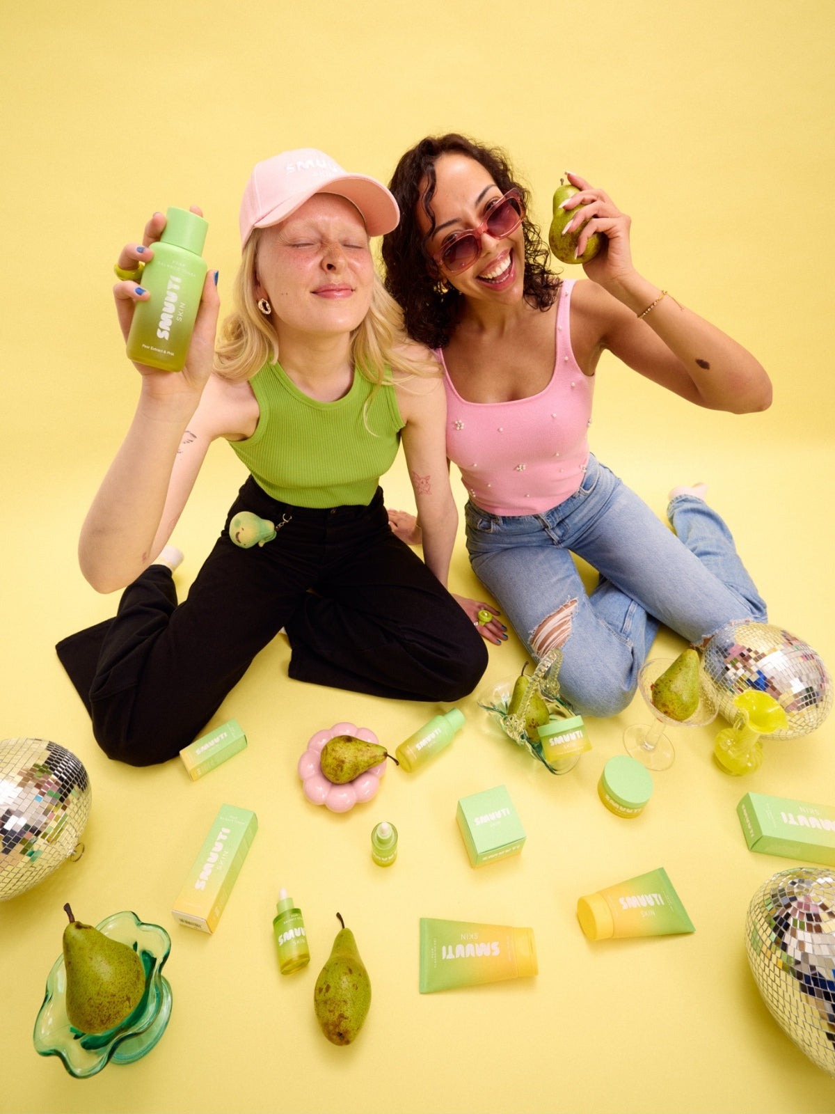 Two women sitting on a yellow floor with pear balance skincare products around them.