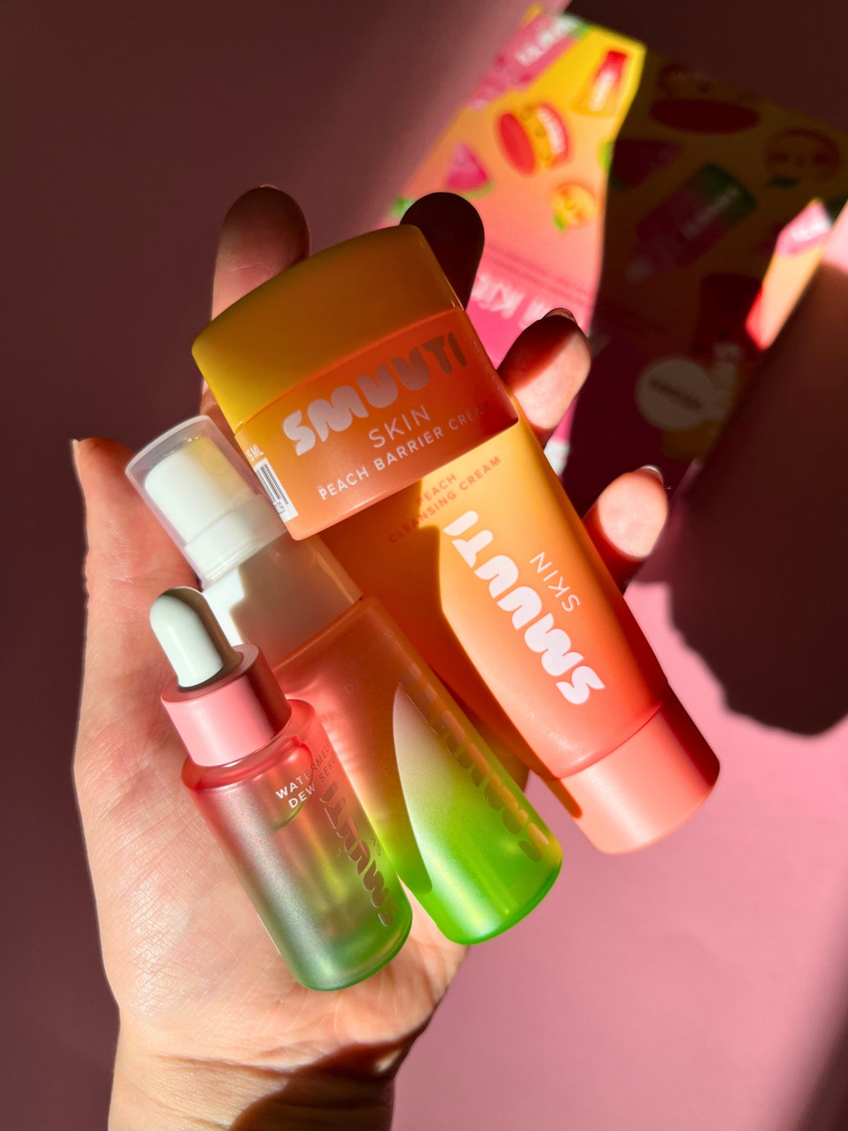 Hand holding travel size Smuuti Skin skincare products with colorful packaging against a pink background