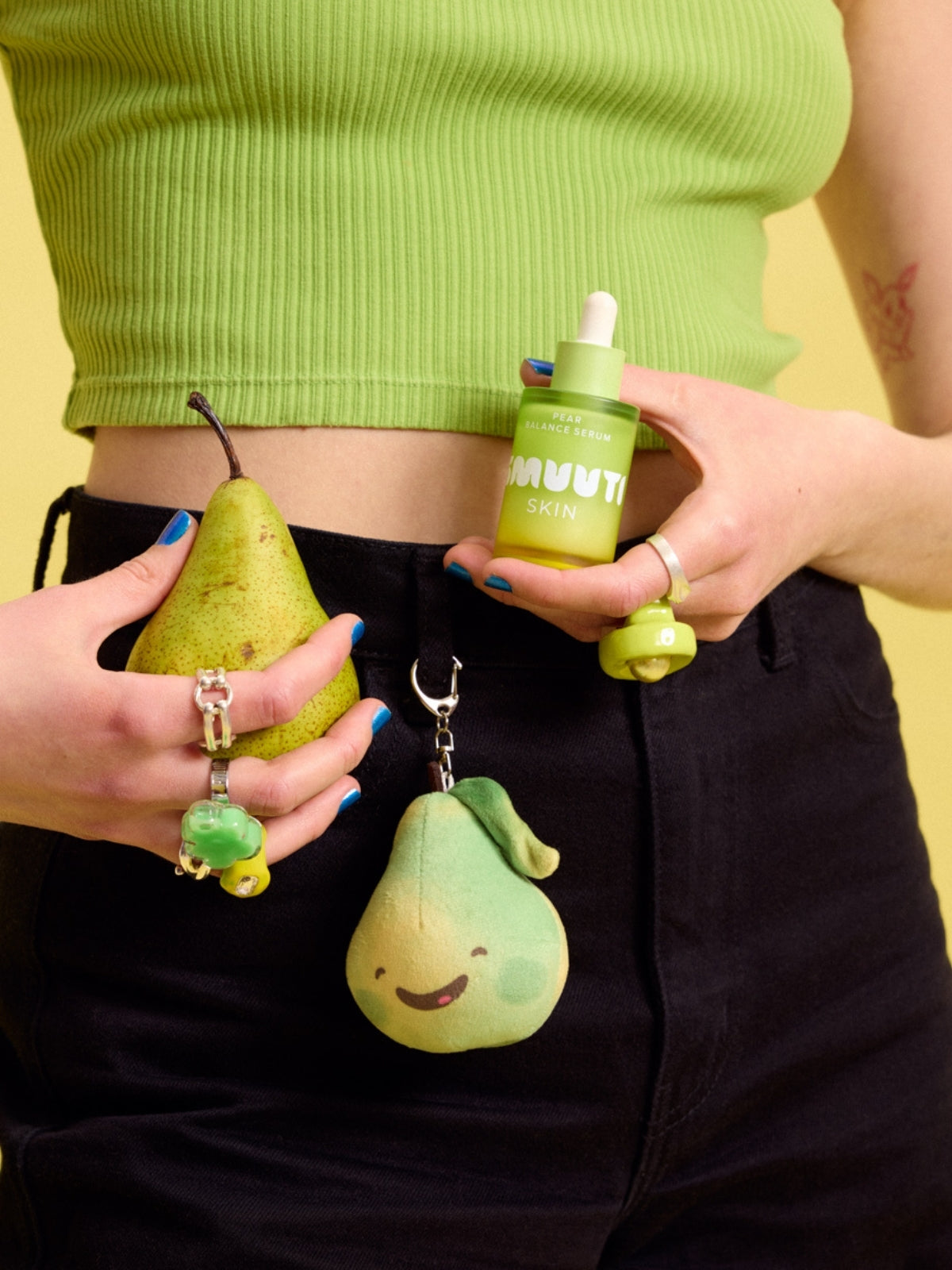Person holding a green pear, Smuuti Skin pear balance serum and a plush toy keychain against a yellow background.