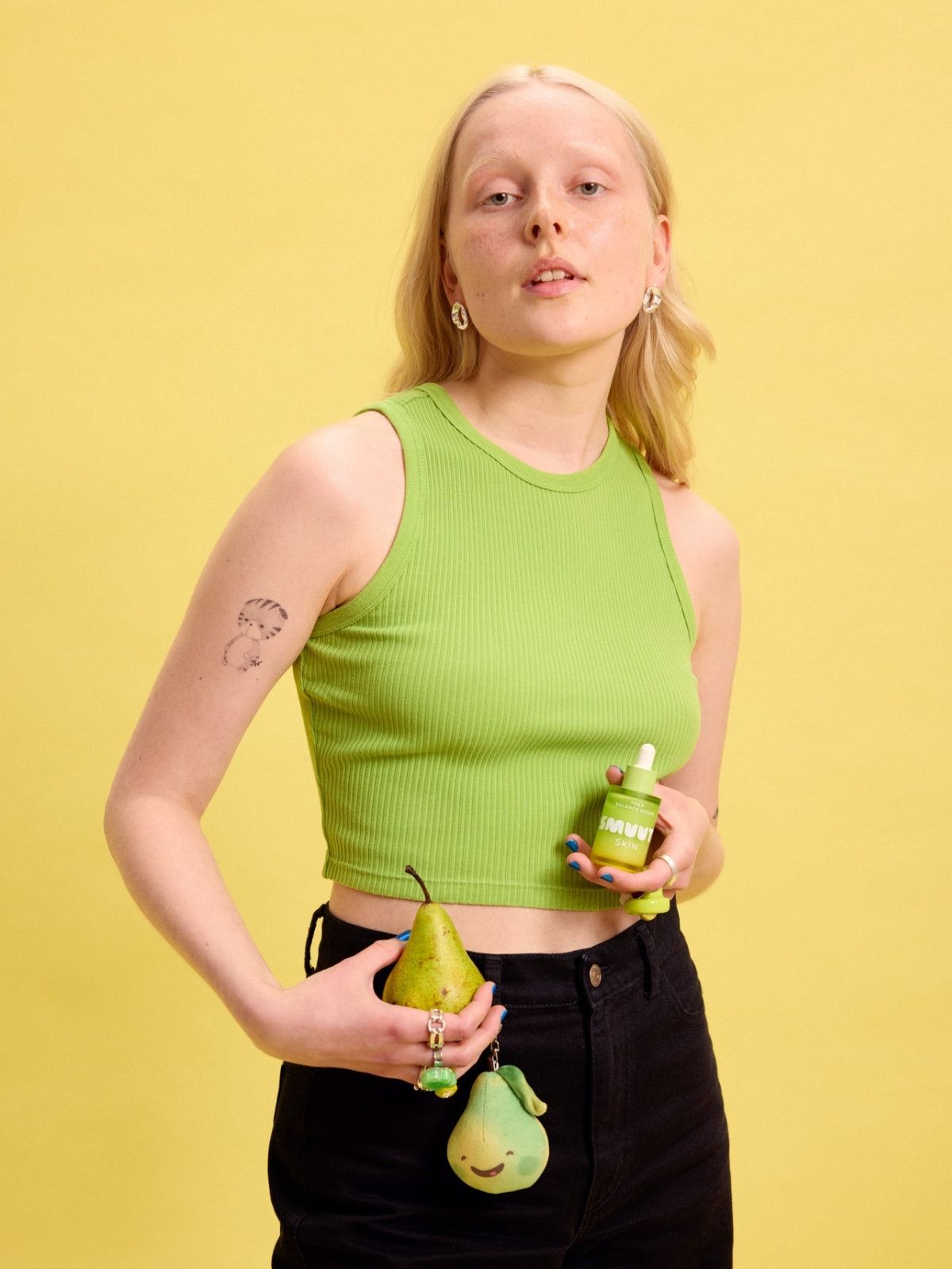 Person wearing a green top holding a green pear, Smuuti Skin pear balance serum and a plush toy keychain against a yellow background.