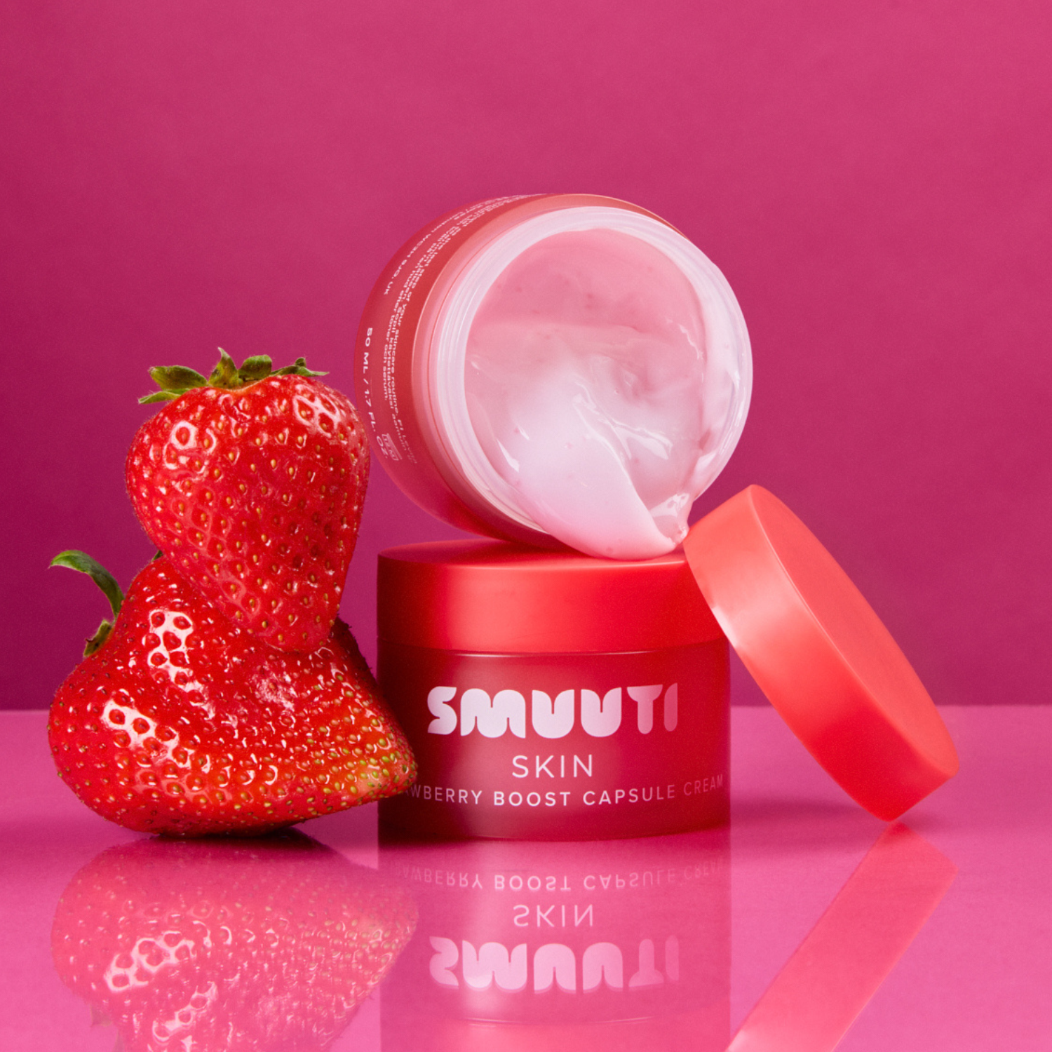 A jar of SMUUTI Skin Strawberry Boost Capsule Cream with its lid off, showing the pink cream inside, styled with two fresh strawberries on a reflective pink surface against a solid pink background.