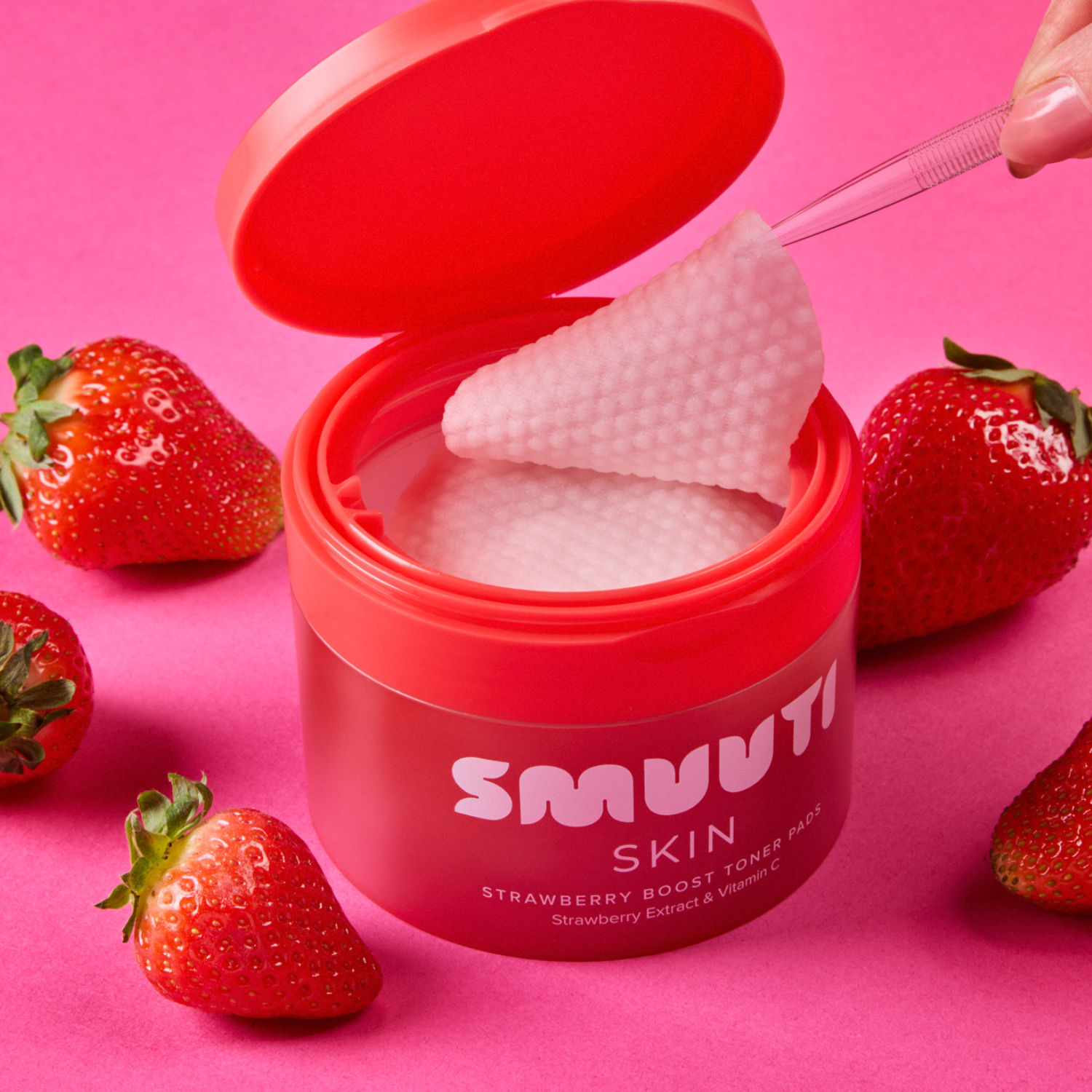 SMUUTI Skin Strawberry Boost Toner Pads shown in a red jar with the lid open, revealing a textured pink pad being lifted by a hand, surrounded by fresh strawberries on a pink background.