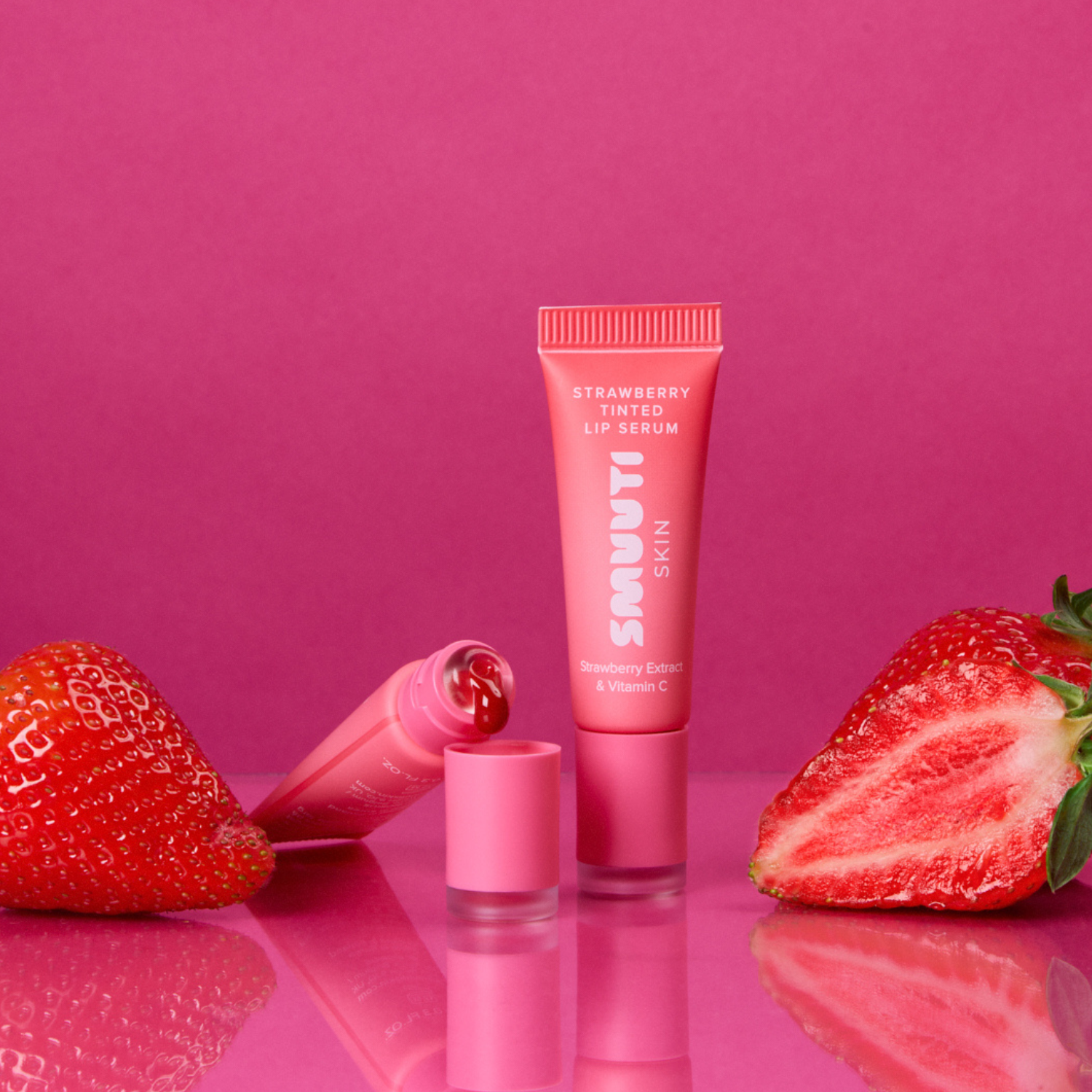 A pink tube of Smuuti Skin Strawberry Tinted Lip Serum stands upright on a glossy pink surface, with the cap beside it and two fresh strawberries placed on either side. The background is a solid pink studio backdrop.