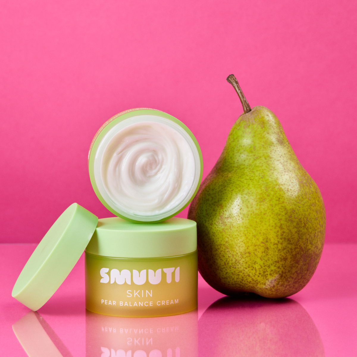An open jar of SMUUTI Skin Pear Balance Cream next to a fresh green pear on a vibrant pink background.