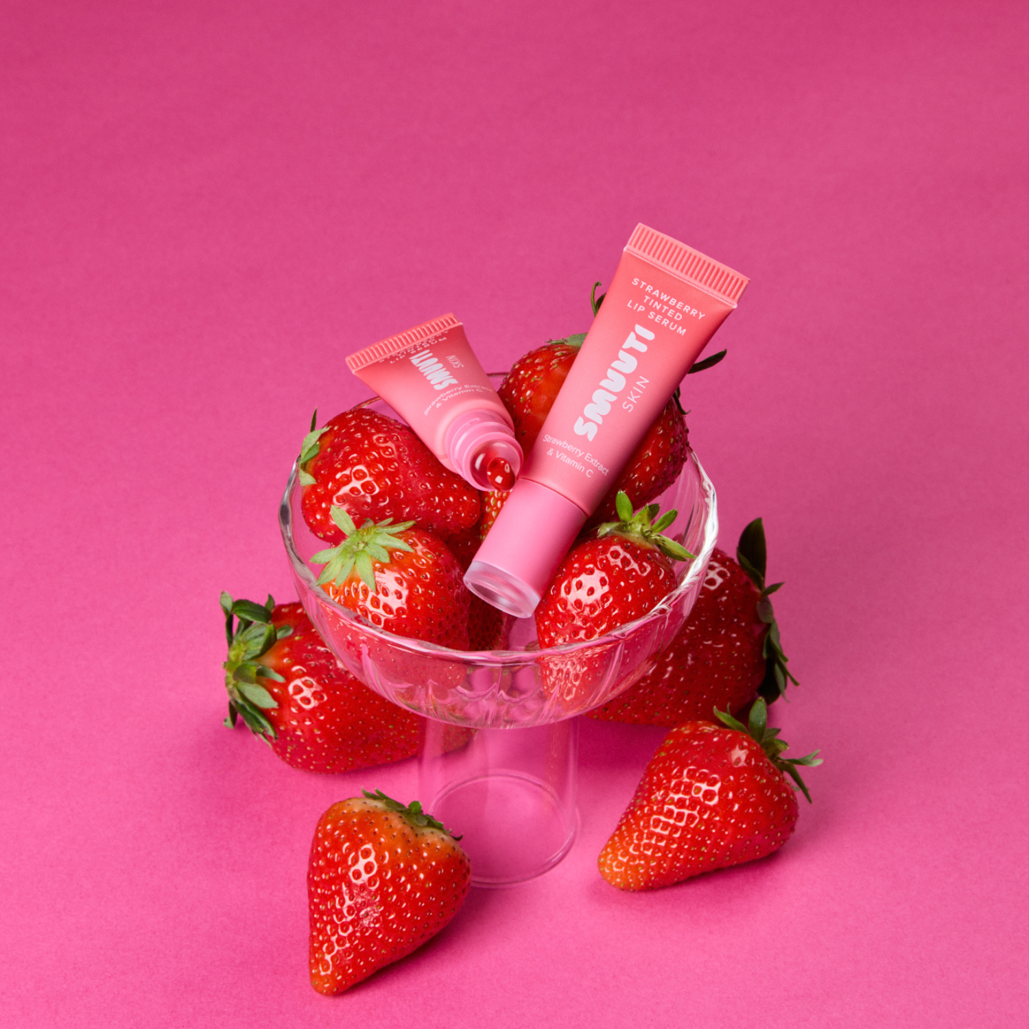 Two strawberry boost tinted lip serum arranged in a clear glass bowl filled with fresh strawberries on a solid pink background.