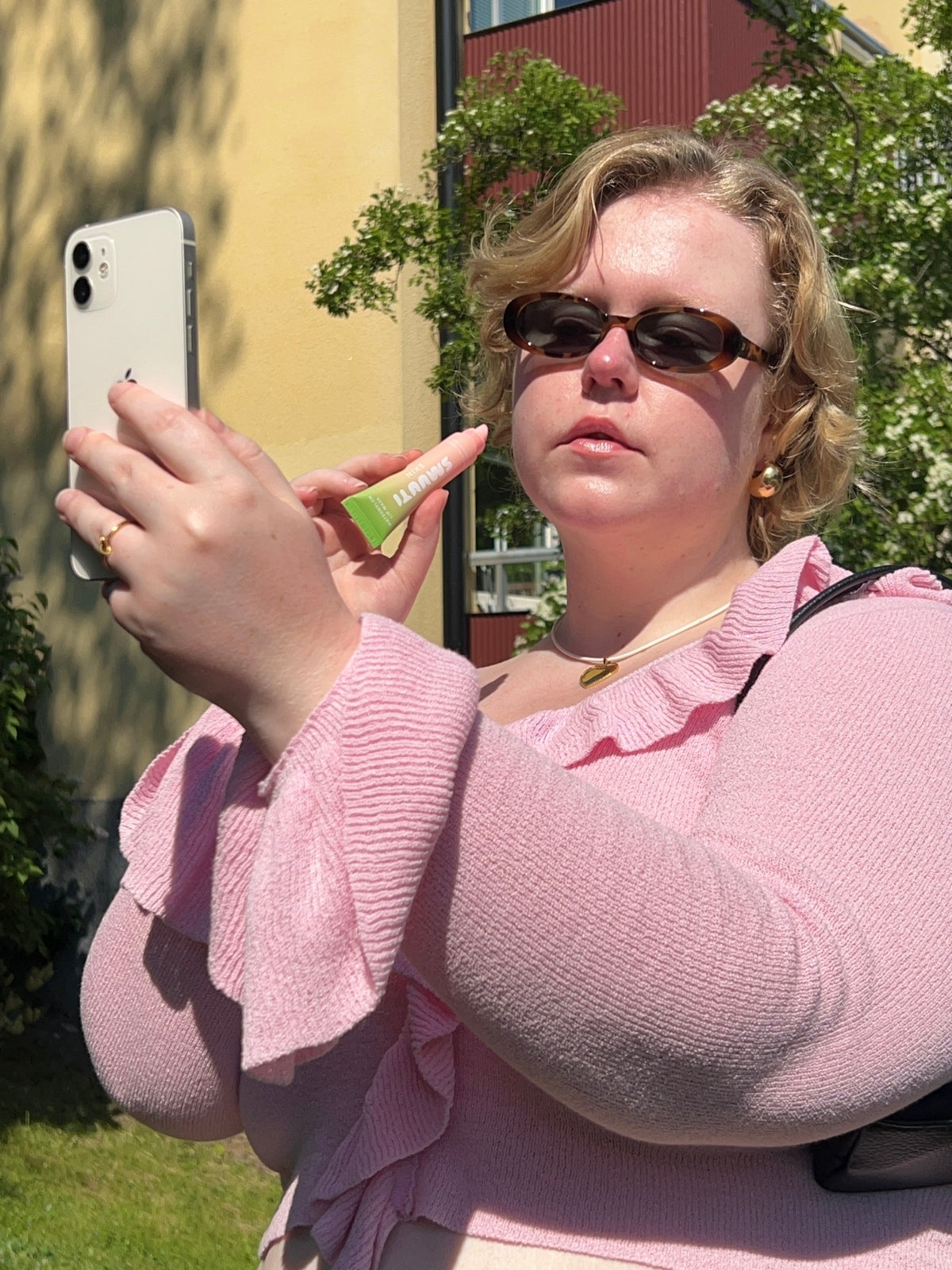 Woman in pink sweater taking a selfie outdoors with a phone. She is holding Smuuti Skin watermelon lip mask
