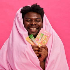 A smiling man wrapped in a pink blanket holds two Smuuti Skin Lip Masks