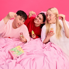 Three people lying in a bed covered with pink sheets, holding Smuuti Skin serums