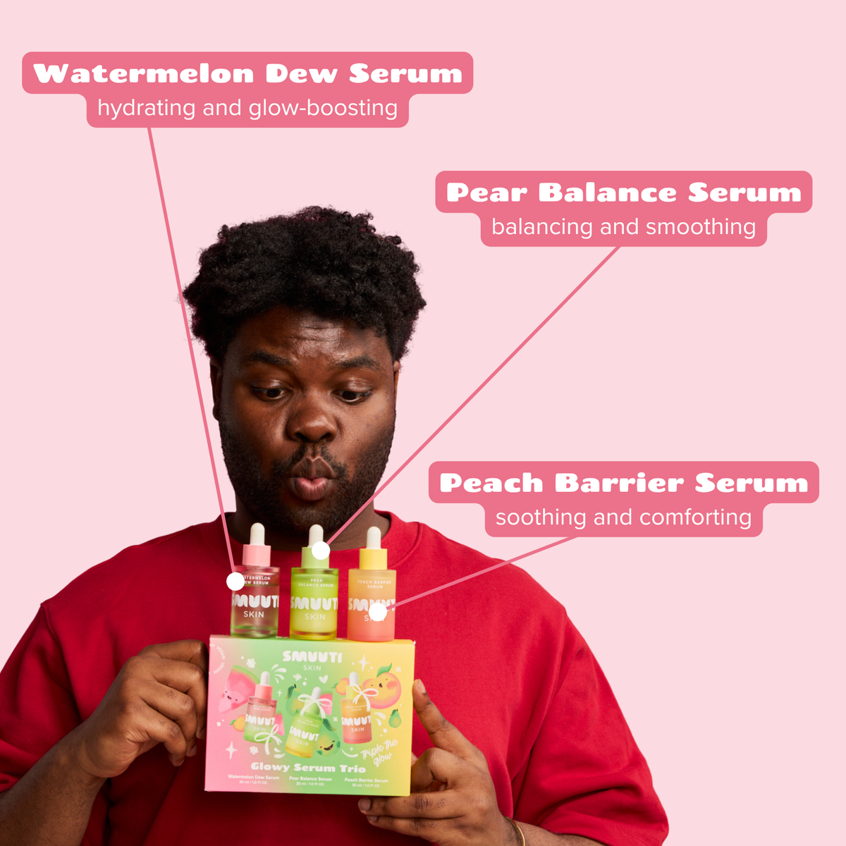A man in a red sweatshirt holds a box of SMUUTI Glow Serum Trio against a solid pink background, with three pink text callouts pointing to the bottles labeled Watermelon Dew Serum, Pear Balance Serum, and Peach Barrier Serum.