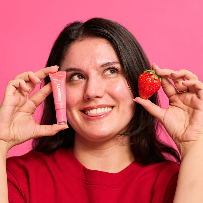 A smiling woman with dark hair holds a pink tube of Glamrus lip product next to a fresh strawberry against a solid pink background.