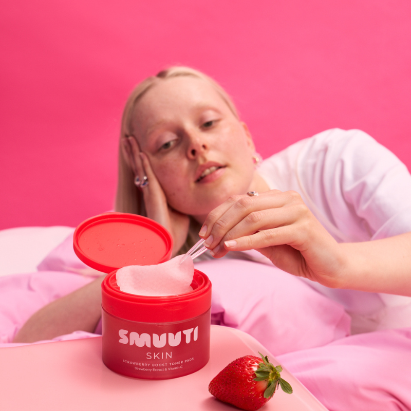 A woman lying on a pink bed uses a tweezers to scoop strawberry boost toner pads. A fresh strawberry sits on the pink bedside table against a solid pink background.