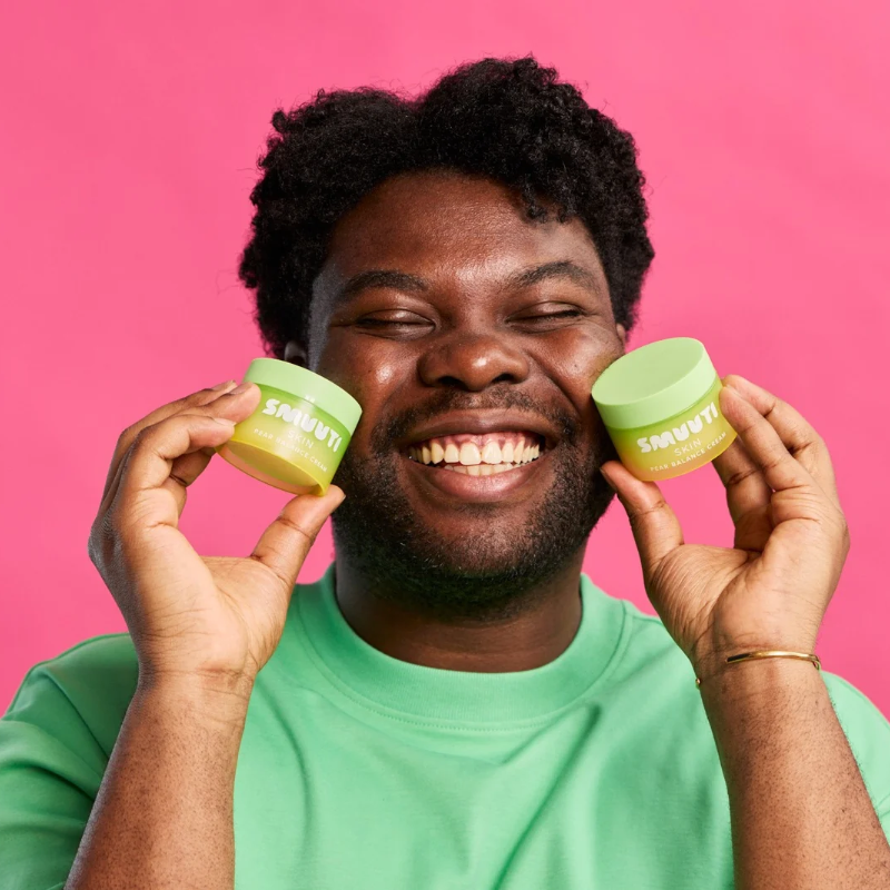 A smiling man with dark curly hair holds two light green jars of Pear Balance Cream