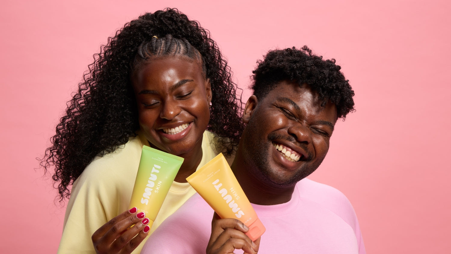 Two people holding colorful Smuuti Skin skincare products against a pink background