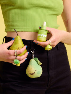 Person holding a green pear,  Smuuti Skin pear balance serum and a plush toy keychain against a yellow background.