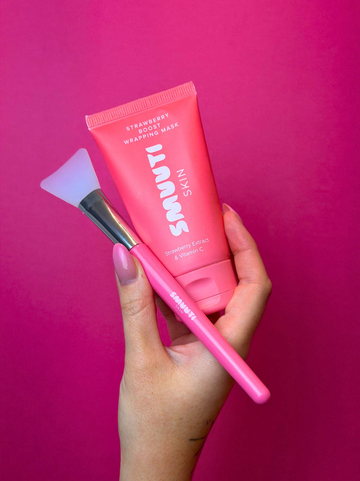 Hand holding a Smuuti Skin wrapping mask product with spatula applicator against a pink background