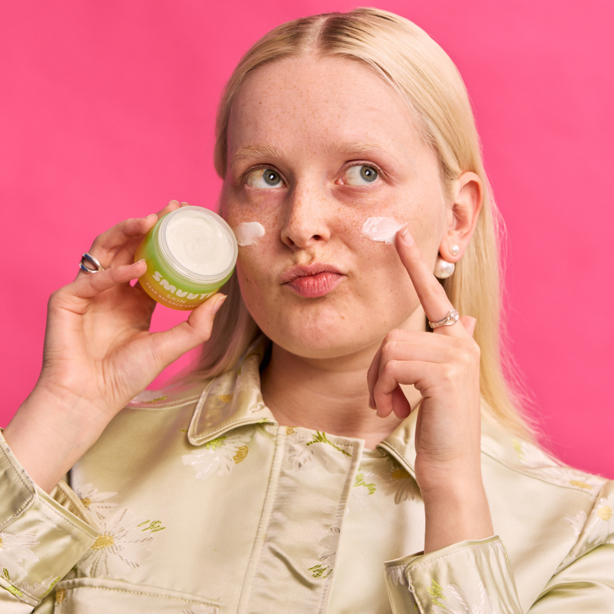 A blonde woman with freckles applies white cream to her cheek while holding a green and white cosmetic jar. She is wearing a light beige floral satin shirt against a solid bright pink background.