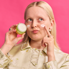A blonde woman with freckles applies white cream to her cheek while holding a green and white cosmetic jar. She is wearing a light beige floral satin shirt against a solid bright pink background.