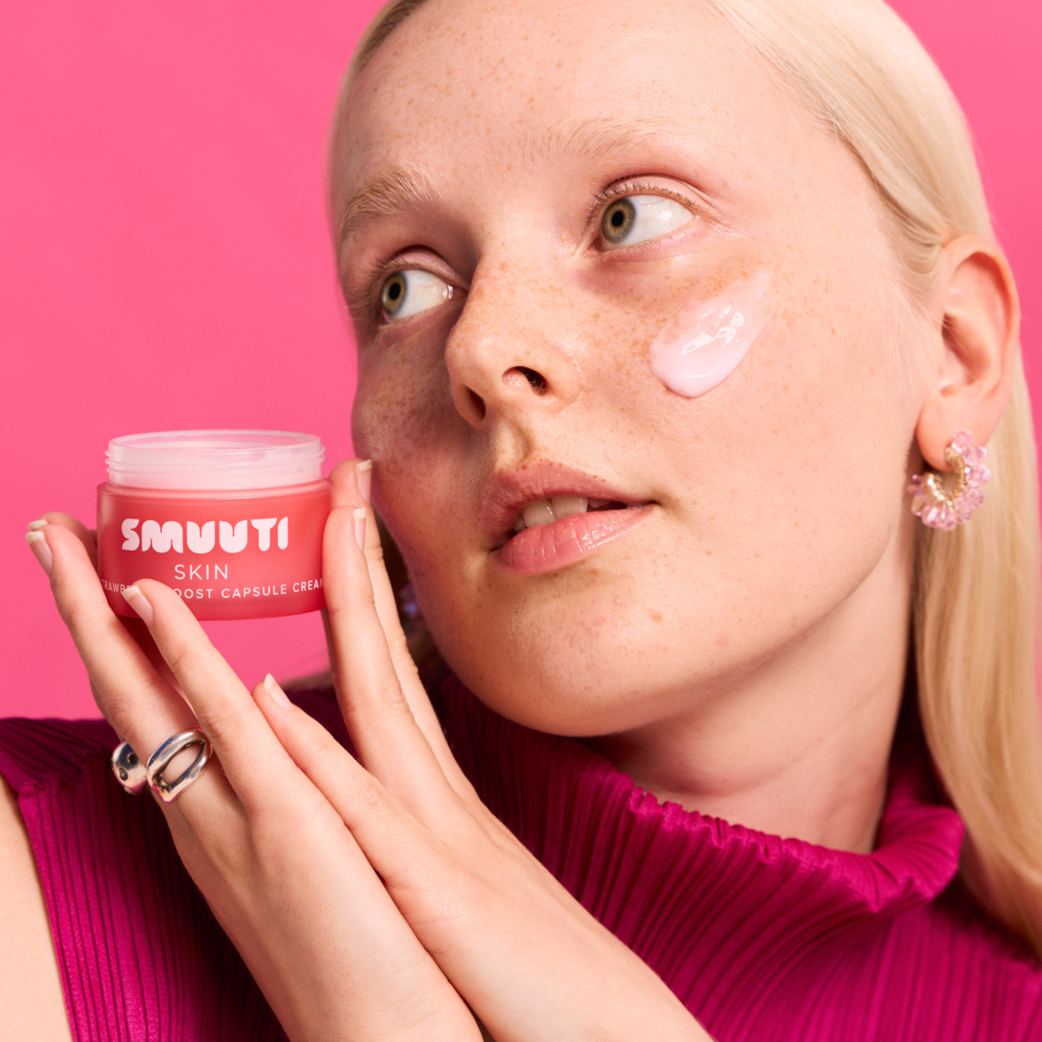 A close-up lifestyle shot of a woman with freckles applying or showing a cream on her cheek, holding a jar of SMUUTI SKIN  Capsule Cream. The background is a solid bright pink, and she is wearing a matching magenta top.